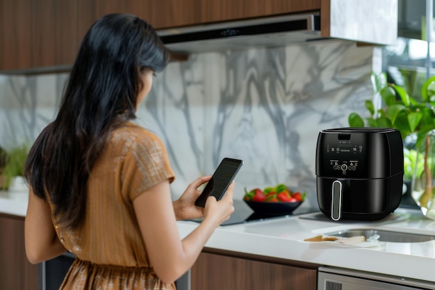 Photo young woman using smartphone to control air fryer in modern kitchen