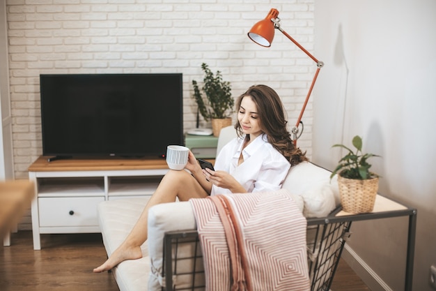 Young woman using smartphone sitting on couch at home. Young person drinks coffe in big mug and serfing internet