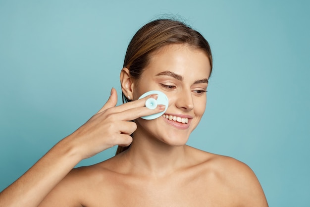 Young woman using using silicone facial cleansing brush on blue wall.