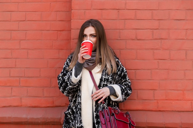 A young woman on a walk in the city drinks coffee from a craft red cup A beautiful blonde woman in a stylish coat stands near a red brick wall on a sunny spring day