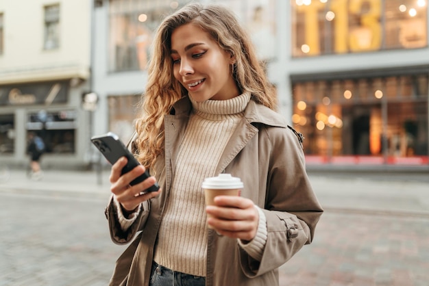 Young woman wearing autumn coat walking with smartphone and coffee cup in a city street