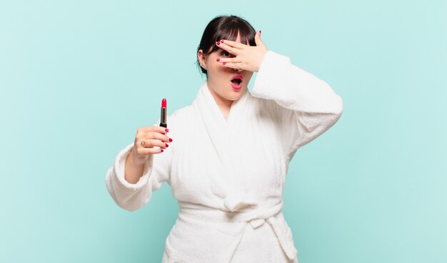 young woman wearing bathrobe looking shocked, scared or terrified, covering face with hand and peeking between fingers