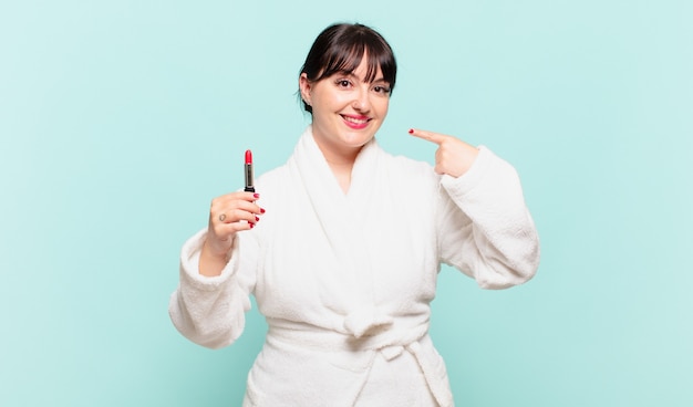 Young woman wearing bathrobe smiling confidently pointing to own broad smile, positive, relaxed, satisfied attitude