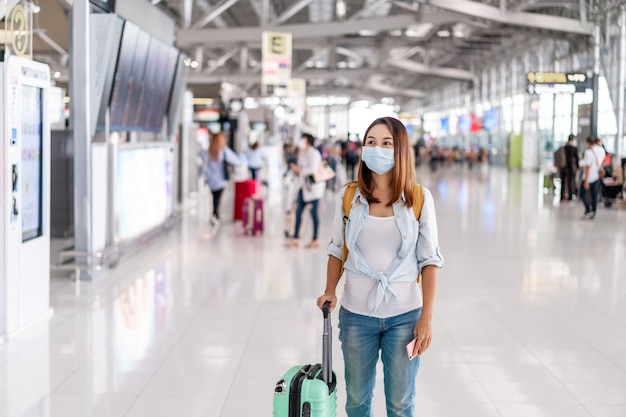 Young woman wearing a face mask and holding a mobile phone is looking for a check-in counter at the airport, New normal lifestyle concept