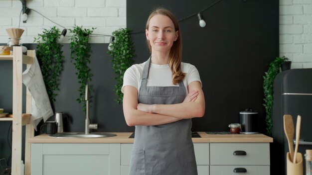 Young woman wearing gray apron smiling and crossing arms standing in her kitchen