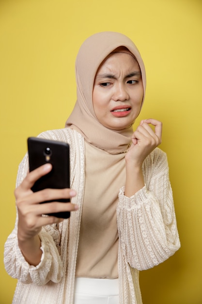 Young woman wearing hijab with an expression disgusted while looking her phone isolated on yellow background