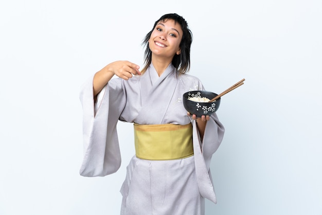 Young woman wearing kimono holding a bowl of noodles on isolated white points finger at you with a confident expression