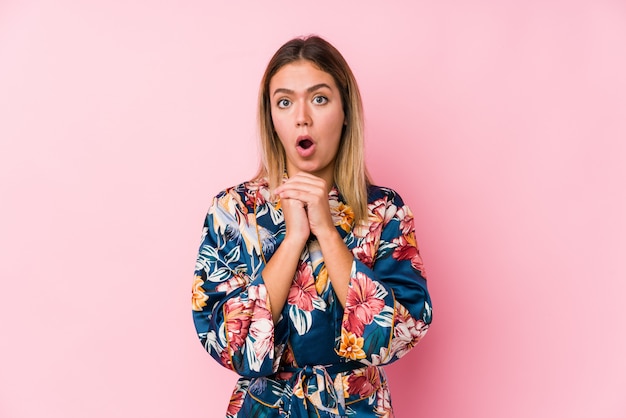 Young woman wearing pajamas praying for luck, amazed and opening mouth looking to front.