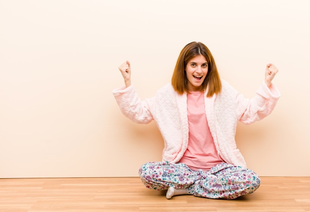 Young woman wearing pajamas sitting at home feeling happy, surprised and proud, shouting and celebrating success with a big smile