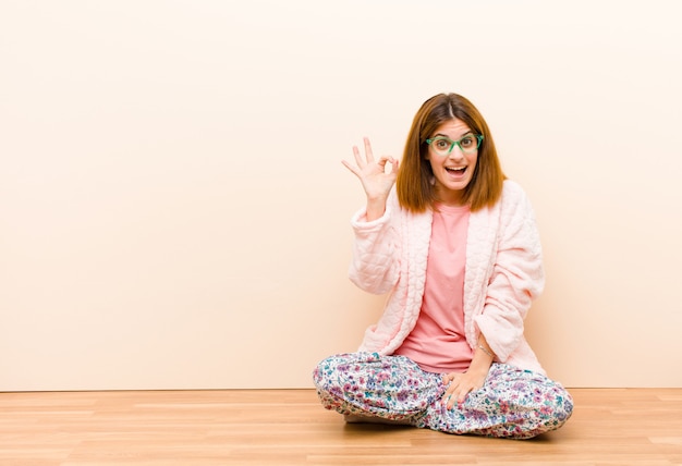 Photo young woman wearing pajamas sitting at home feeling successful and satisfied, smiling with mouth wide open, making okay sign with hand