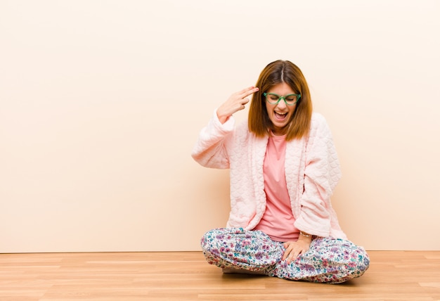 Photo young woman wearing pajamas sitting at home looking unhappy and stressed, suicide gesture making gun sign with hand, pointing to head