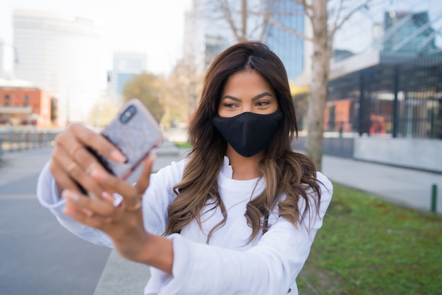 young woman wearing protective mask and taking selfies with her mophile phone while standing outdoors. Urban concept.