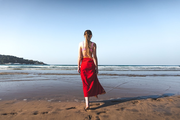 Young woman wearing a red towel and a pink bikini walking at the water edge on the beach