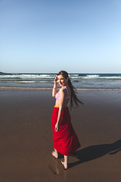 Young woman wearing a red towel and a pink bikini walking at the water edge on the beach