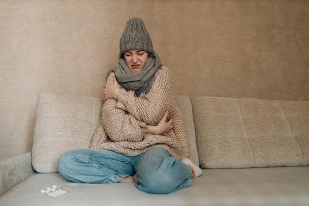 Young woman wearing scarf and wool hat has flu ill sick disease cold at home indoor on sofa