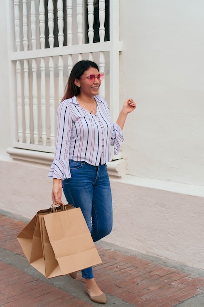 Young woman wearing sunglasses carrying shopping bags while standing by wall