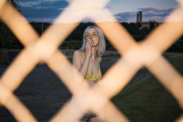 Photo young woman wearing sunglasses smoking while standing on field seen through fence at dusk