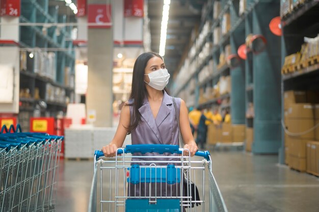 A young woman wearing a surgical mask with a trolley in shopping mall, covid-19 and pandemic concept