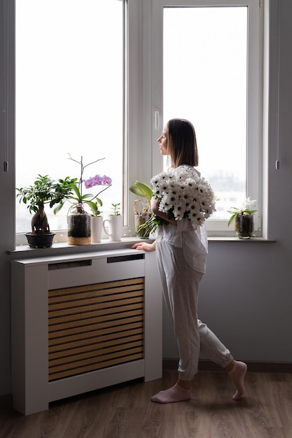 Young woman wearing white casual clothes looking out window holding a bouquet of white flowers waiting for spring or summer to come