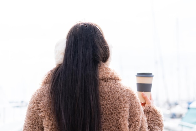 Young woman wearing winter muffs and holding take away coffee at outdoors in back position