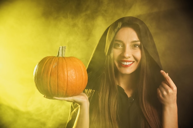 Young woman wearing witch costume with pumpkin in smoke cloud on dark background Halloween party