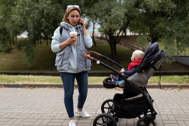 Young woman while walking with her baby and pram talking on a mobile phone and drinking coffee in a