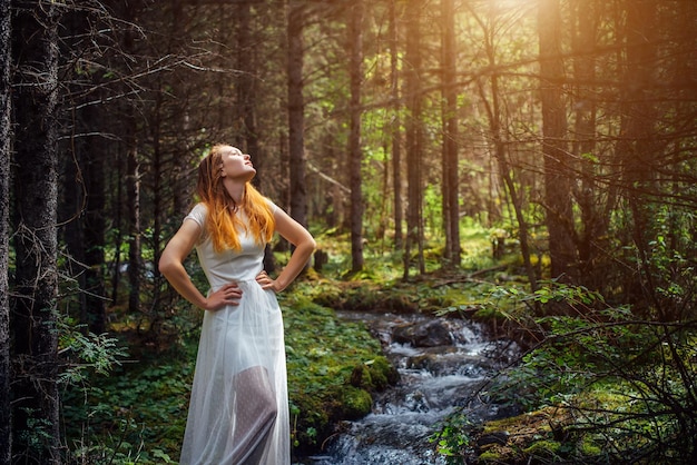 Young woman in white dress raised her face towards the sun Dreamy pretty girl on the background of green forest and mountain stream