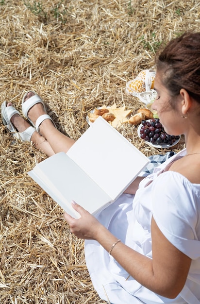 Young woman in white dress sitting on haystack in harvested field reading blank book book mockup