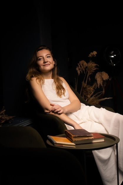 A young woman in a white dress smiles and sits relaxed on an armchair in a dark room