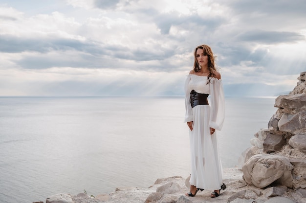 A young woman in a white dress stands barefoot on a cliff face in full height The dress flutters in the wind A sacred glow breaks through the clouds The girl looks like an angel