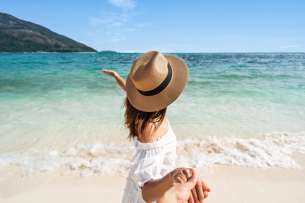 Young woman in white dress and straw hat holding partner's hand at the beach