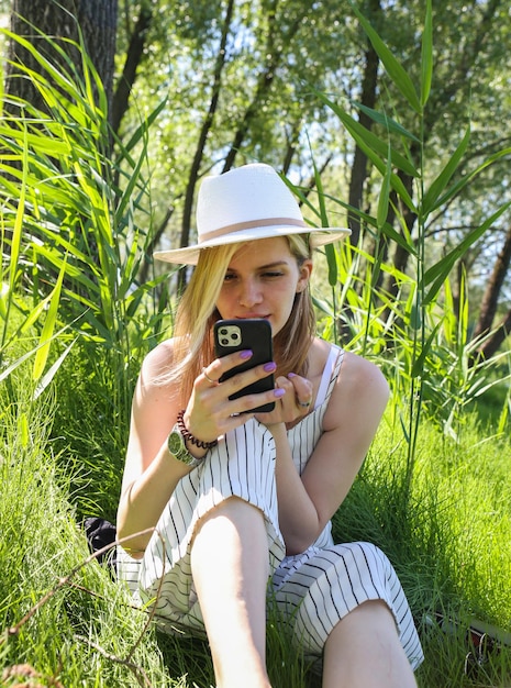 a young woman in a white hat is sitting in the grass with a phone in her hand