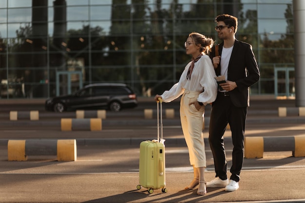 Young woman in white outfit and handsome brunette man in black suit pose near airport Charming blonde girl in blouse and beige pants holds yellow luggage