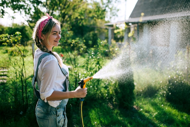 a young woman in a white shirt and denim overalls watering the lawn