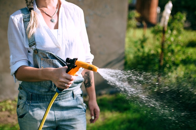 a young woman in a white shirt and denim overalls watering the lawn