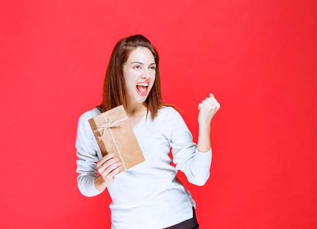 Young woman in white shirt holding a cardboard gift box and showing positive hand sign