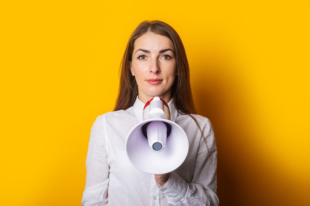 Young woman in a white shirt holds a megaphone in her hands on a yellow background