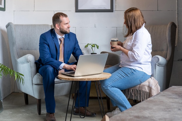 A young woman in a white shirt and jeans holding a mug of coffee and a middleaged Arab man in a business suit talking at a cafe table with a laptop Business people discussing new project