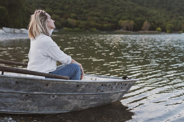 Young woman in a white shirt and jeans sits on a boat and looks at a mountain lake