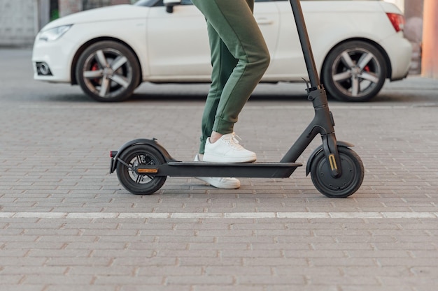 Young woman in white sneakers is standing on electric scooter in front of the white car