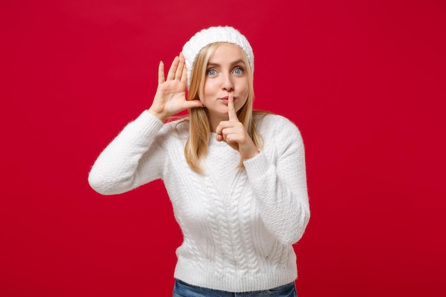 Young woman in white sweater, hat isolated on red background. Healthy fashion lifestyle cold season concept. Mock up copy space. Saying hush be quiet with finger on lips shhh gesture, try to hear you.