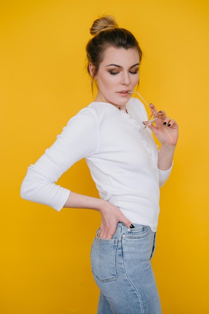 A young woman in a white sweatshirt and blue jeans stands in the studio against a yellow background