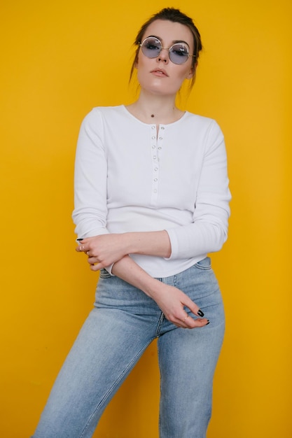 A young woman in a white sweatshirt and blue jeans stands in the studio against a yellow background