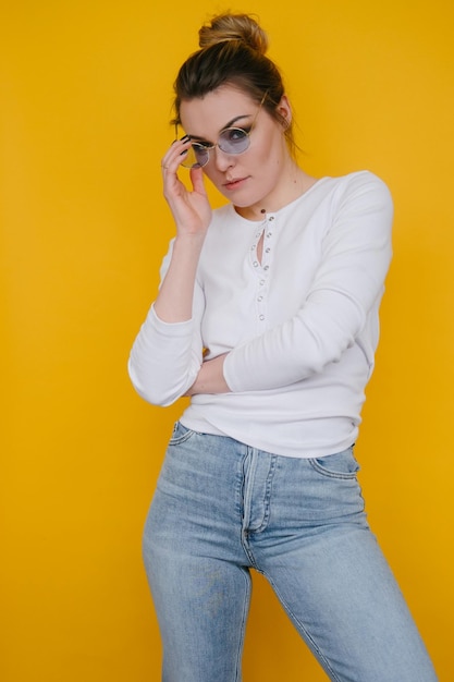 A young woman in a white sweatshirt and blue jeans stands in the studio against a yellow background