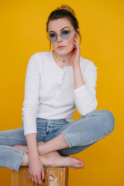 A young woman in a white sweatshirt and blue jeans stands in the studio against a yellow background