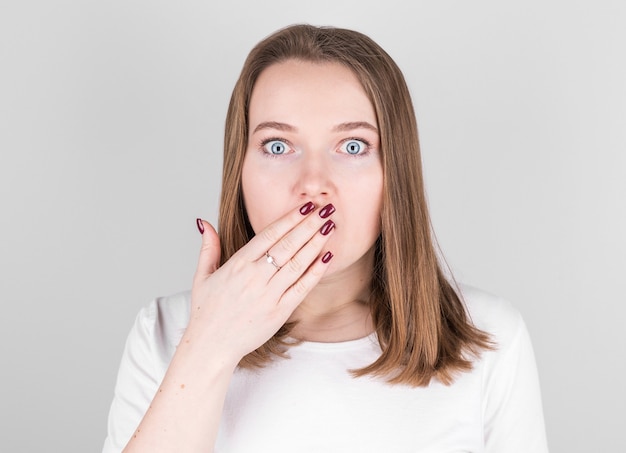 Photo young woman in a white t-shirt stands against a gray wall and covers her mouth