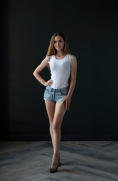 young woman in a white tank top and denim shorts on a dark background, full length portrait