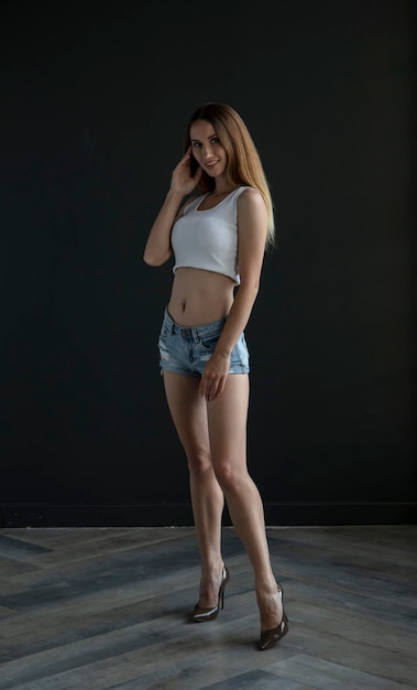 young woman in a white tank top and denim shorts on a dark background, full length portrait