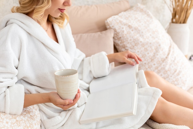 Young woman in white terry robe is drinking coffee and reading magazine or book in bedroom