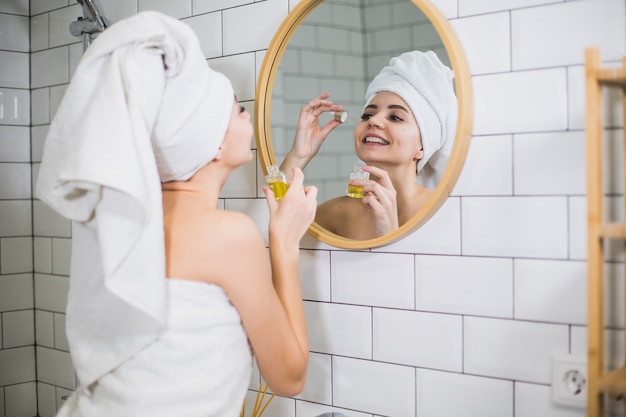 Young woman in white towel apply moisturising oil on face skin. Self care, beauty and skin care concept.